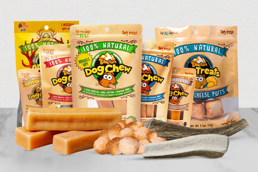A group of Tinker Treats dog treats and packaging displayed on a light gray surface. Visible products include multiple bags of dog treats and several pieces of the treats arranged in front, including rectangular cheese puffs, round pieces, and a chew on a stick.